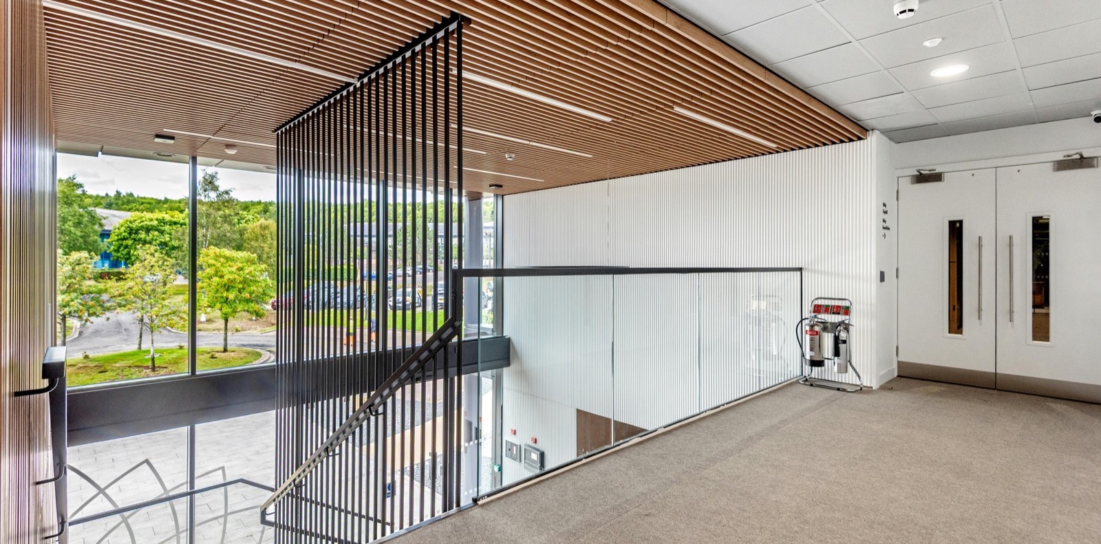First-floor landing with timber-slatted ceiling, glass balustrade and full-height glazing overlooking the car park.